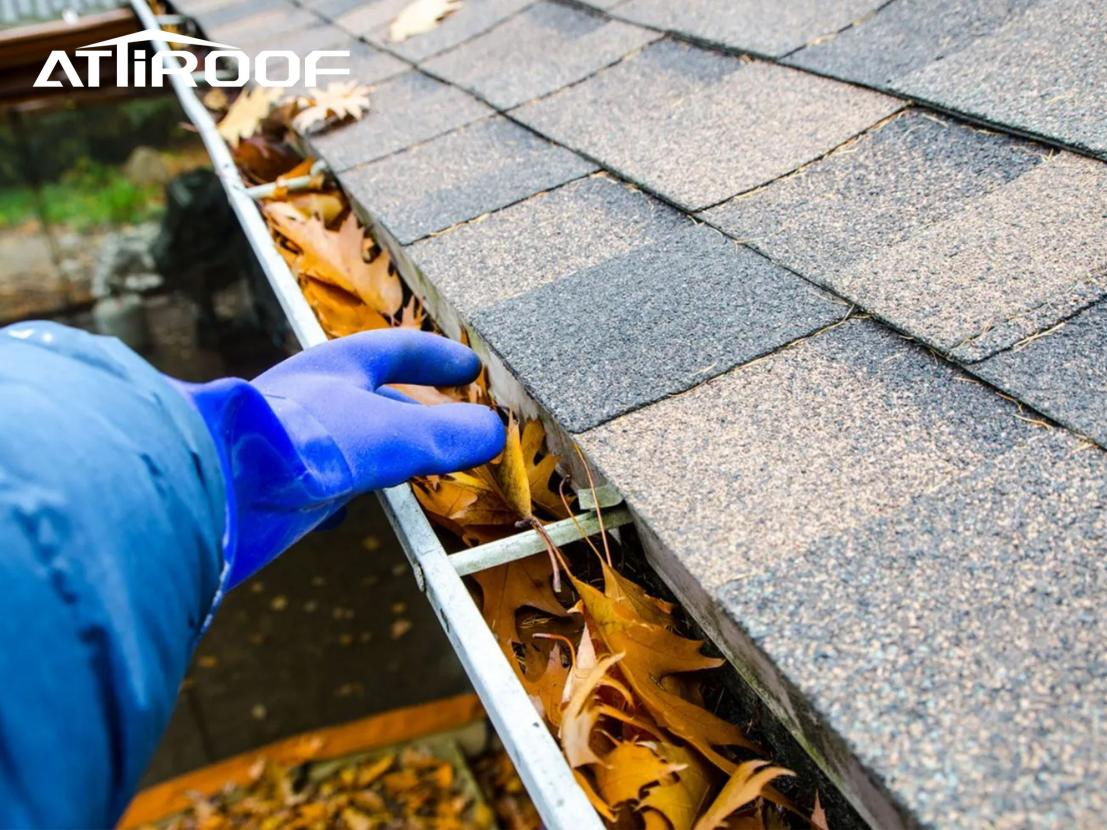 A person wearing a blue coat and gloves is clearing yellow and brown fallen leaves from the roof drainage ditch.