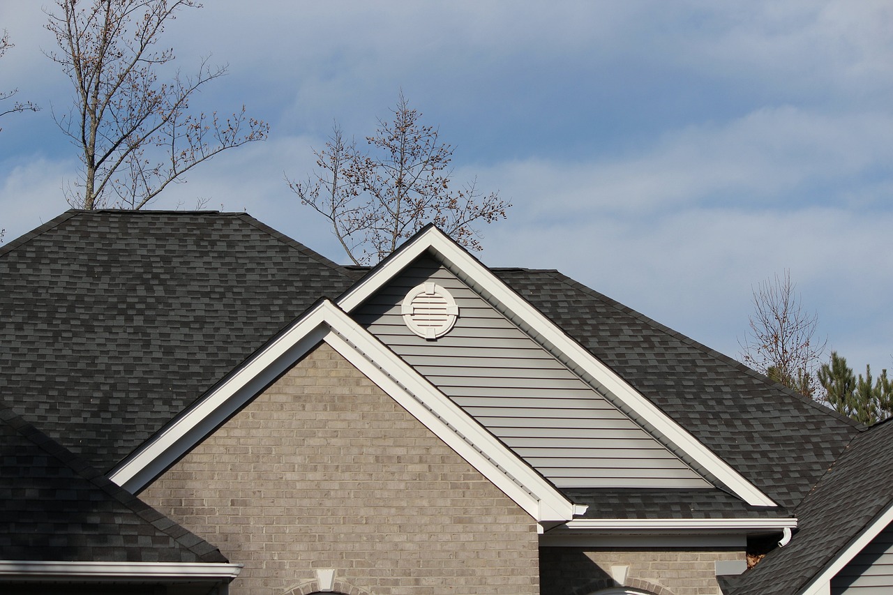 A house with a dark gray asphalt tile roof and white eaves, located under deciduous trees and blue sky and white clouds.